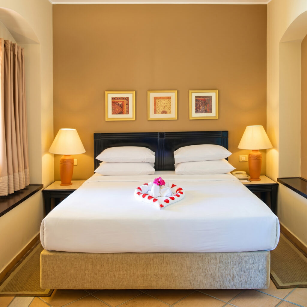 Luxury Bedroom Suite Interior Perfectly symmetrical luxury hotel bed with a decorative towel heart and flower petals, illuminated by warm bedside lamps at Steigenberger Coraya Beach.